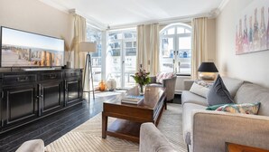 A modern living room with a flat-screen TV, dark wood cabinets, a grey sofa, and a large window with curtains. - Residence Bismarck, Whg. 9 - Residence Bismarck (Sylt)