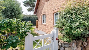 Une maison en briques avec un portail blanc, un jardin où poussent différentes espèces de plantes et un mur en pierre.