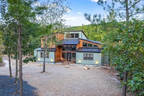 A modern house with a large deck, surrounded by trees and a gravel driveway. - Unwind, Play, and Make Memories — Your Perfect Getaway Awaits! Beautiful creek! (Hochatown)