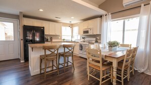 A modern kitchen with wooden cabinets, a central island with bar stools, a dining table with chairs, and a large window with curtains.