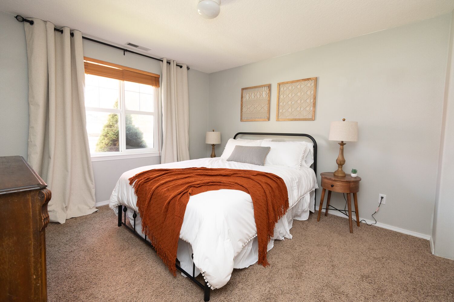 A bedroom with a bed, two bedside lamps, a window with curtains, a small wooden nightstand, and a wall-mounted lamp.
