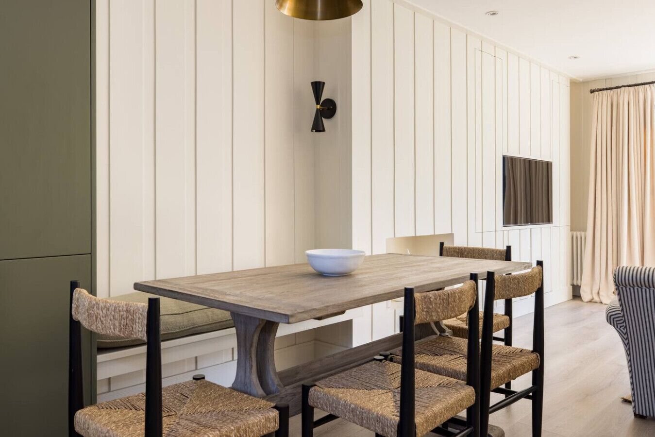A dining area with a wooden table and woven chairs, a wall-mounted light fixture, and a television on the wall.