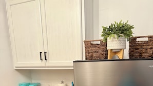 Una cocina con gabinetes blancos, un refrigerador de acero inoxidable, una encimera de madera, una cafetera verde azulada, un recipiente blanco para azúcar y una cesta de mimbre con una planta.