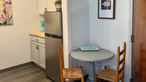 Una cocina compacta con armarios blancos, un refrigerador de acero inoxidable, una pequeña mesa de comedor con dos sillas de madera y un cuadro en la pared.