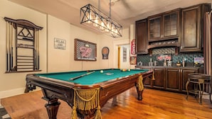 A pool table in a room with wooden floors, a bar area, and a kitchen with dark cabinets. - Majestic & Relaxing Home w/ 3 Beds Pool Table Bar! Sleeps 8 (Dubuque)