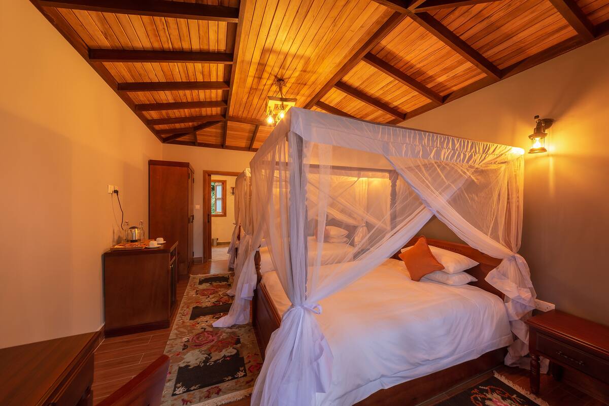 A bedroom with a four-poster bed, wooden ceiling beams, a nightstand, and a night lamp.