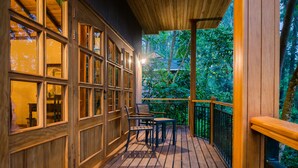 A wooden deck with a table and chairs, surrounded by glass doors and windows, overlooking a lush garden. - Chimpundu Lodge (Fort Portal)