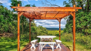 Una pérgola de madera con una mesa de picnic y bancos en un entorno natural.