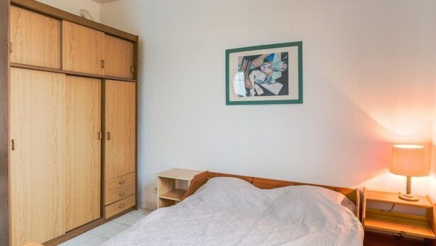 Ein Schlafzimmer mit Bett, Holzschrank, Nachttisch, Lampe und einem gerahmten Bild an der Wand.