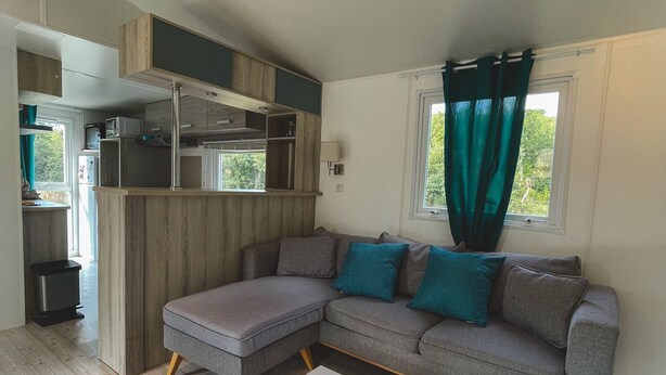A compact living space with a kitchenette, a grey sectional sofa, a white coffee table, and a window with curtains. - Charming mobile home in the heart of nature (Vidauban)