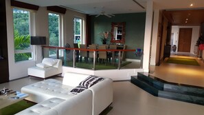 Uma sala de estar moderna com um sofá branco, uma área de jantar com guarda-corpo de vidro e vista para o verde lá fora.