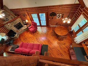 Interior de una cabaña con chimenea de piedra, un sofá rosa, una mesa redonda de madera y una escalera que conduce a un balcón.