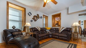 A living room with a brown leather sectional sofa, two side chairs, a wooden coffee table, and a large window with blinds.