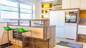 A modern kitchen with a wooden island, two green bar stools, a built-in oven, and a window with blinds. - Apartment Sabine by Interhome (Oberperfuss)