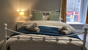 Un lit bien fait, avec un cadre en métal blanc, une couverture bleue et deux serviettes enroulées. Une table de chevet avec une lampe et une boîte de mouchoirs se trouve à côté du lit. Un tableau représentant des fleurs de cerisier est accroché au-dessus du lit.