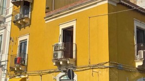 Un edificio giallo con una persiana chiusa, un piccolo balcone con ringhiera in metallo e un'auto parcheggiata davanti.