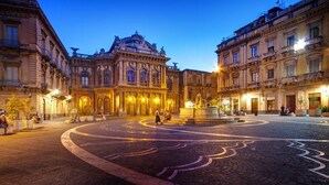 Una piazza storica con edifici decorati, una fontana al centro e pavimentazione in sampietrini.
