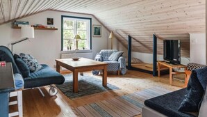 A cozy attic room with a sloped ceiling, wooden flooring, a sofa, a small table, a TV, and a window with a view of greenery.
