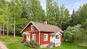 A red wooden house with a white porch, surrounded by lush green trees.