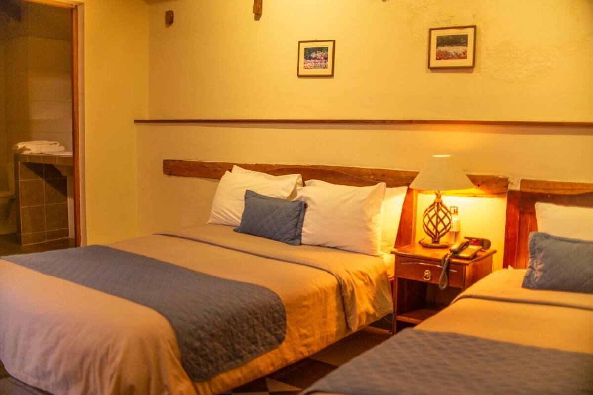 A hotel room with two beds, a wooden headboard, a nightstand with a lamp, and a shower area.