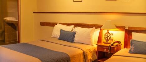 Habitación de hotel con dos camas, cabecero de madera, mesita de noche con lámpara y zona de ducha.