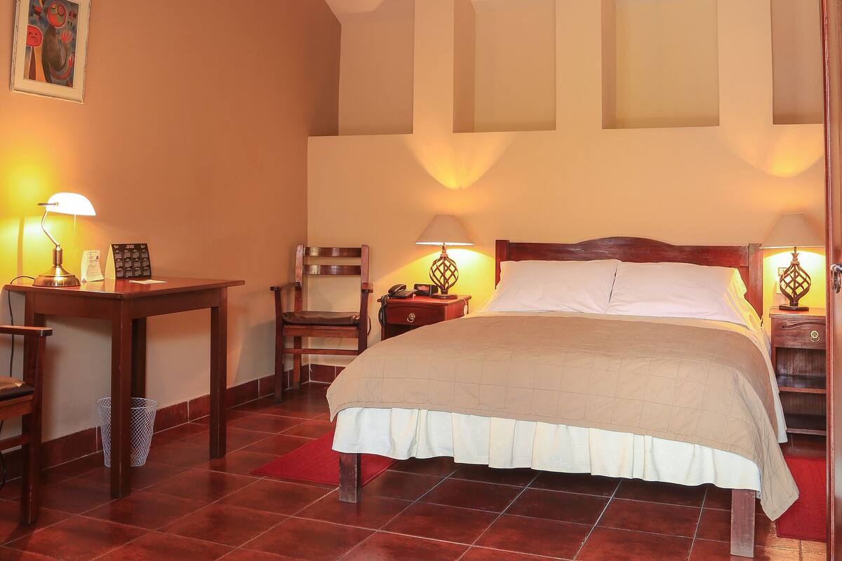 A bedroom with a bed, wooden headboard, a desk with a lamp, a chair, and a red rug.