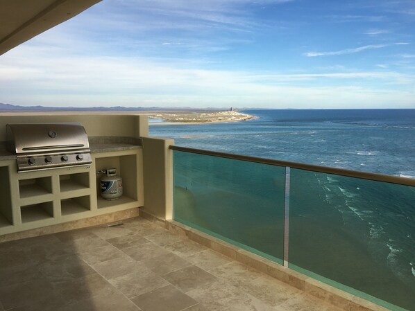A balcony with a barbecue grill and a gas canister, overlooking a coastal area with a clear view of the sea and distant land.