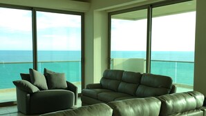 Un soggiorno ampio con un grande divano grigio e una poltrona più piccola, ampie finestre con vista sull'oceano e un balcone con ringhiera.