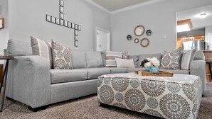 Ein Wohnzimmer mit grauem Sofa, einem gemusterten Hocker und Wanddekoration.