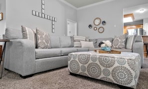 A living room with a gray sofa, patterned ottoman, and decorative wall art.