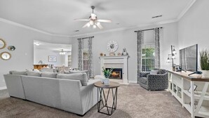 Ein Wohnzimmer mit Kamin, einer Couch, einem gemusterten Sessel, einem Fernsehschrank und einem Deckenventilator.