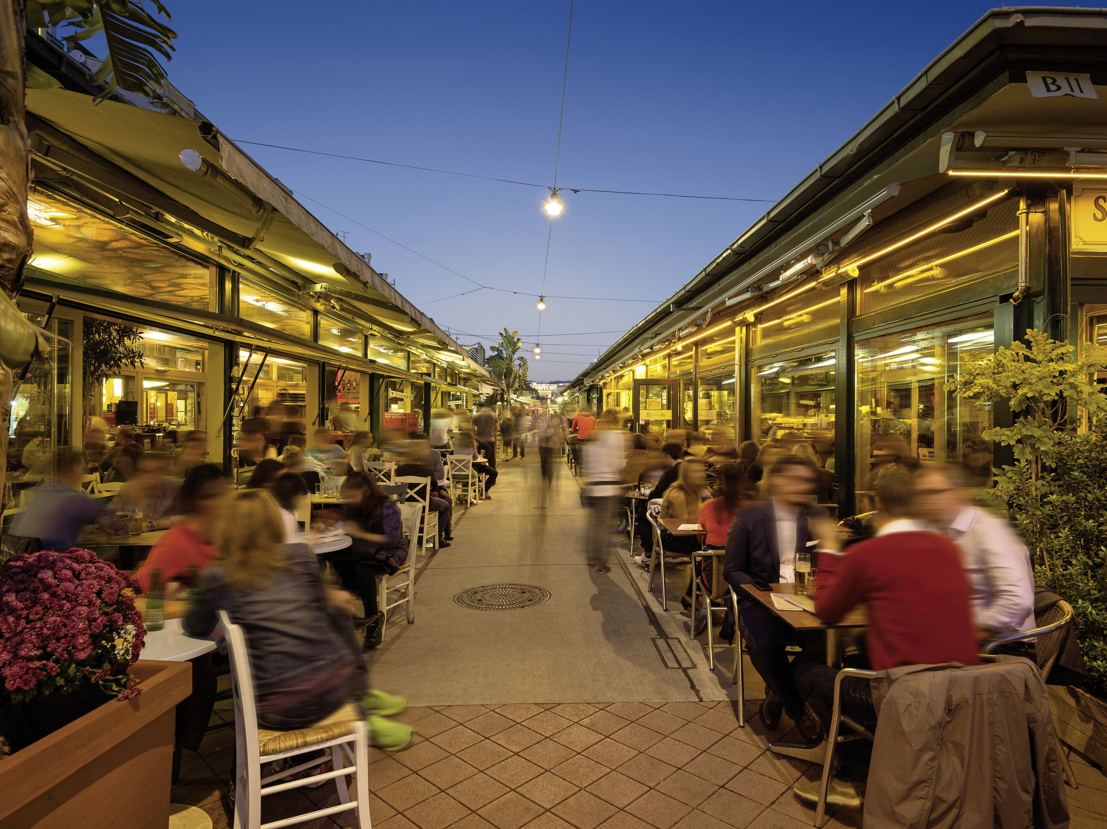 Un marché animé en plein air, avec des tables et des chaises pour s’asseoir et manger, entouré de bâtiments portant des enseignes.