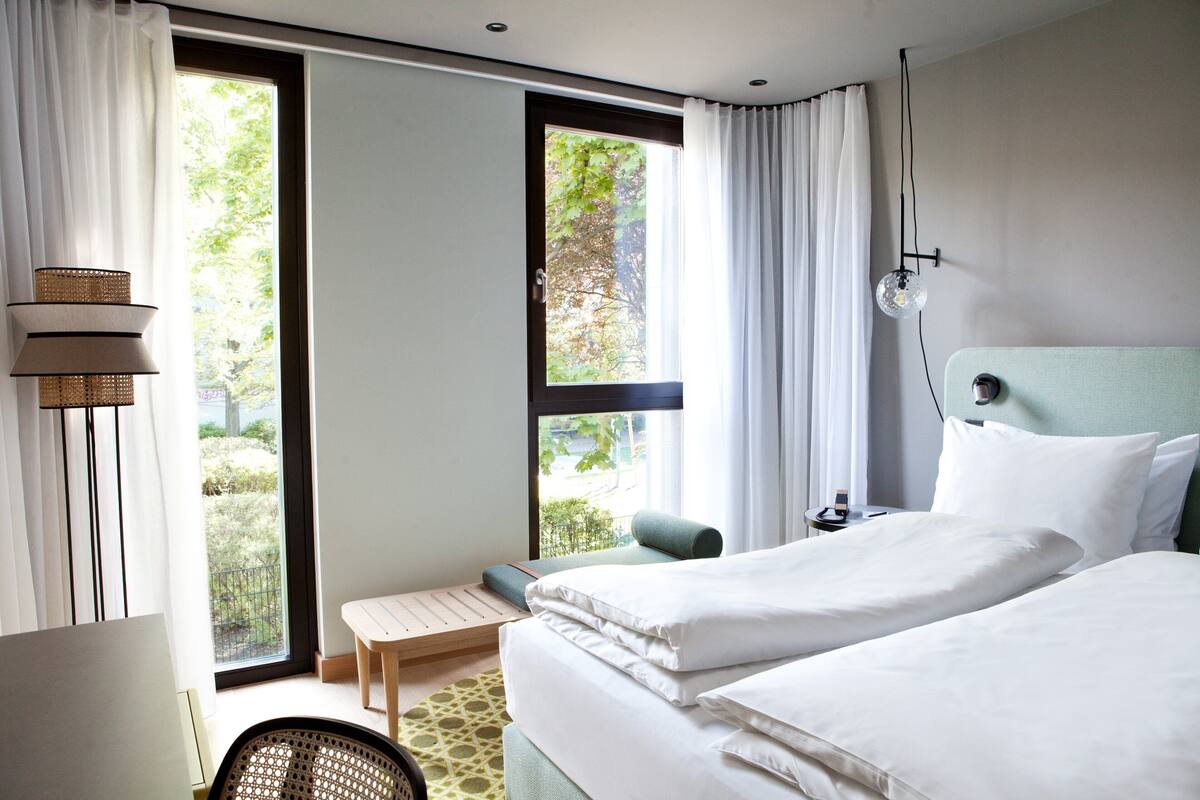 A hotel room with two beds, a small table, a chair, a lamp, and a view of greenery outside.