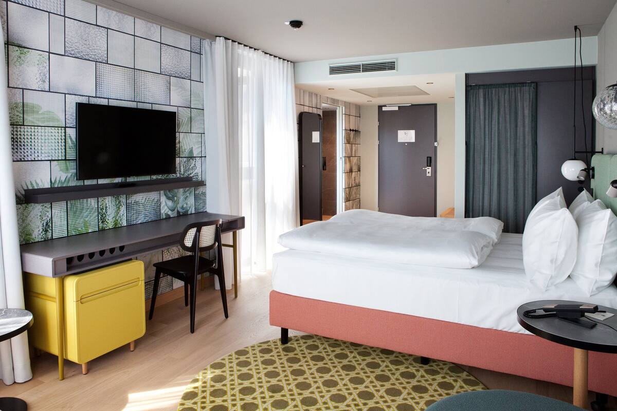 A hotel room with a bed, a desk, a chair, a TV, and a small table.