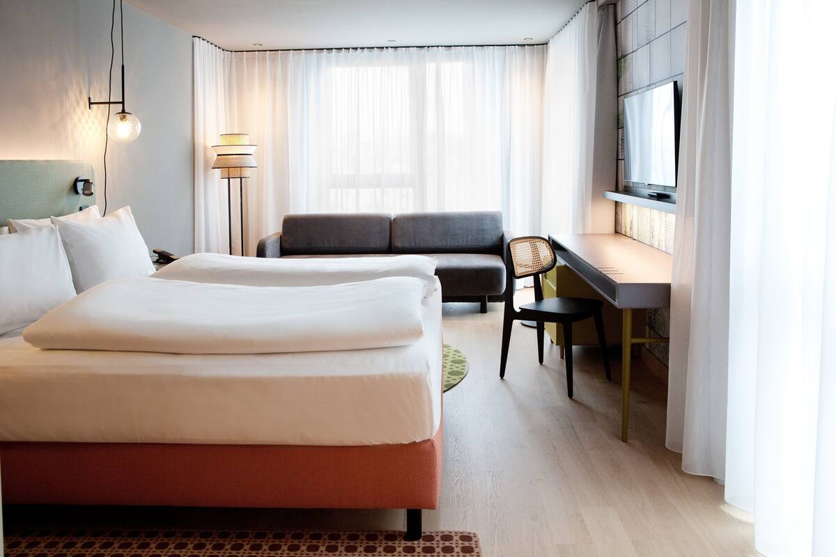 A hotel room with two beds, a desk, a chair, a TV, and a lamp.