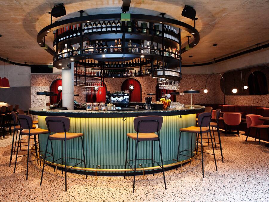 Zona de bar con una barra circular, varios taburetes y una estantería para vinos.