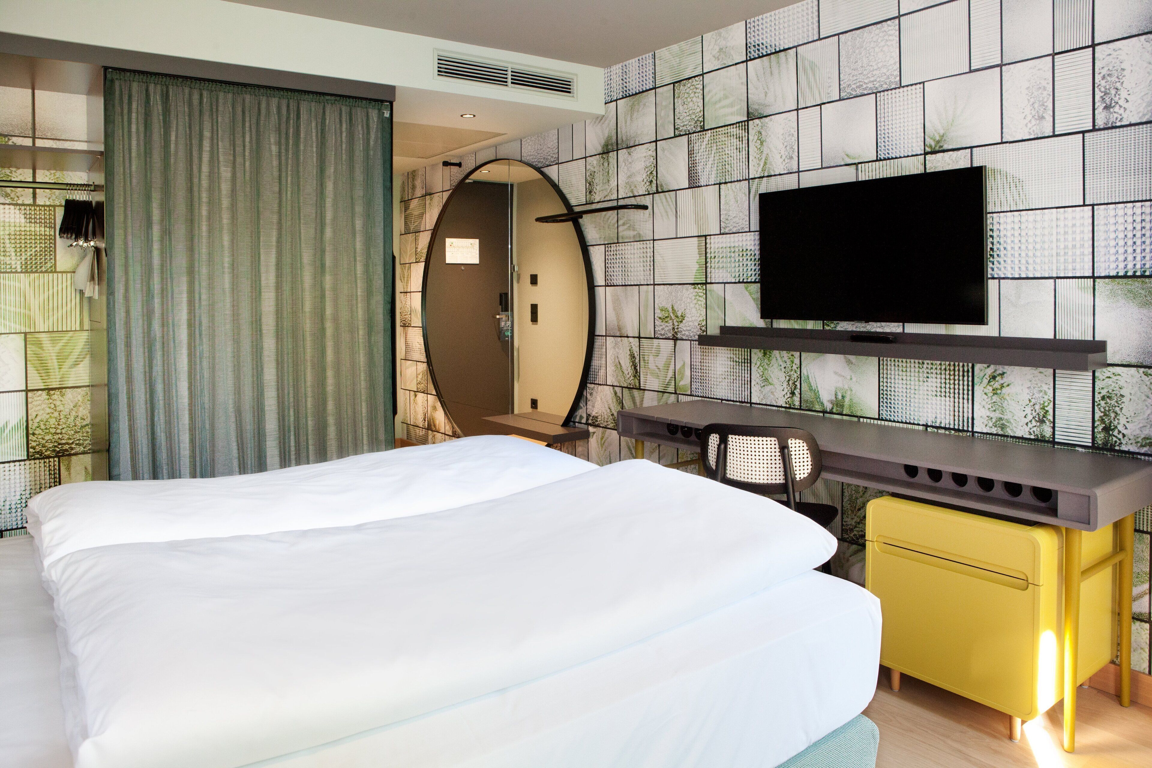 Hotellihuone, jossa on suuri sänky, työpöytä tuolilla, seinälle asennettu taulutelevisio ja peili.