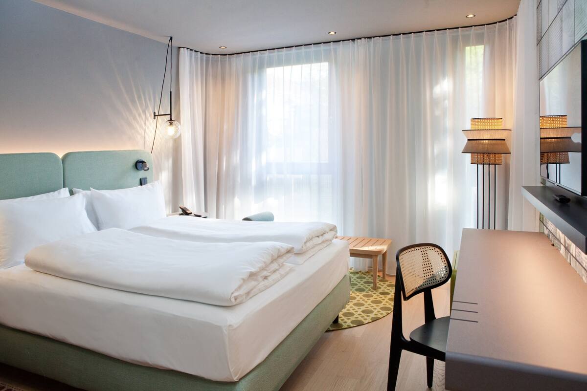 A hotel room with a bed, a desk, a chair, a lamp, and a window with curtains.