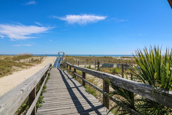 A wooden boardwalk leading to a beach with dunes and a clear sky.