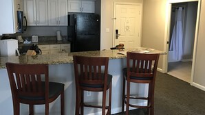 A compact kitchen with white cabinets, a black refrigerator, and a granite countertop. There are three wooden bar stools and a small island.
