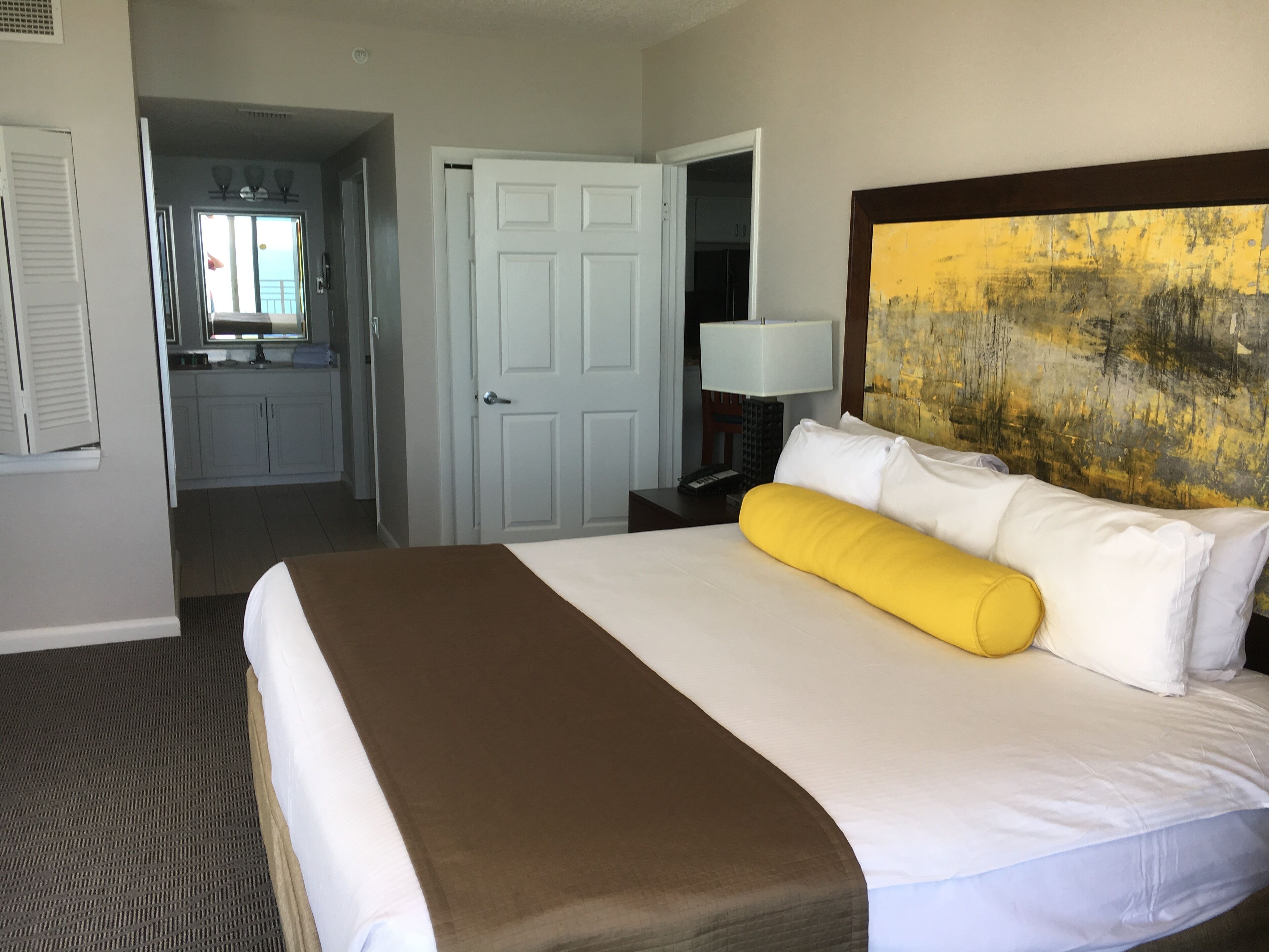 Hotelkamer met een groot bed, een geel kussen, een schilderij aan de muur en uitzicht op de gang en badkamer.