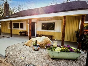 A house with a porch, a large rock, a cat statue, and a flower bed.