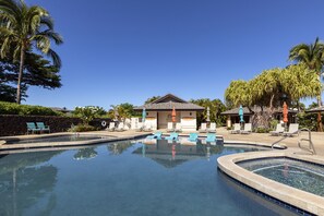 Une piscine entourée de palmiers et d’un bâtiment à toit de tuiles, avec des chaises longues et des parasols à proximité.