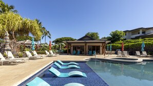 A swimming pool with lounge chairs and umbrellas, surrounded by palm trees and a building with a covered area.