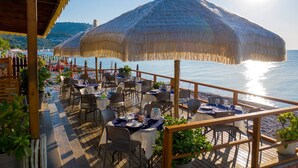 Un espace de restauration en plein air, avec des tables et des chaises, de grands parasols en chaume et une vue sur la mer.