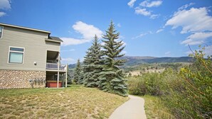 A two-story building with a stone facade, a balcony, and a pathway leading to a large evergreen tree. - Close to 5 Ski Resorts - On Free Bus Route - Blue River in Backyard + Hot Tub (Silverthorne)