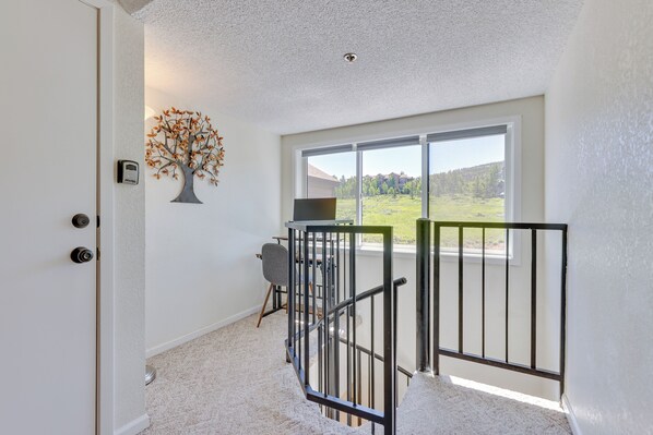 A room with a white door, a tree wall art, a desk with a chair, a window with a view of greenery, and a black railing.