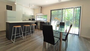 A modern kitchen with a central island, bar stools, a dining table with chairs, and a large sliding glass door leading to a green outdoor area. - New, Modern Property built in 2021 * Close to Town (Glenroy)