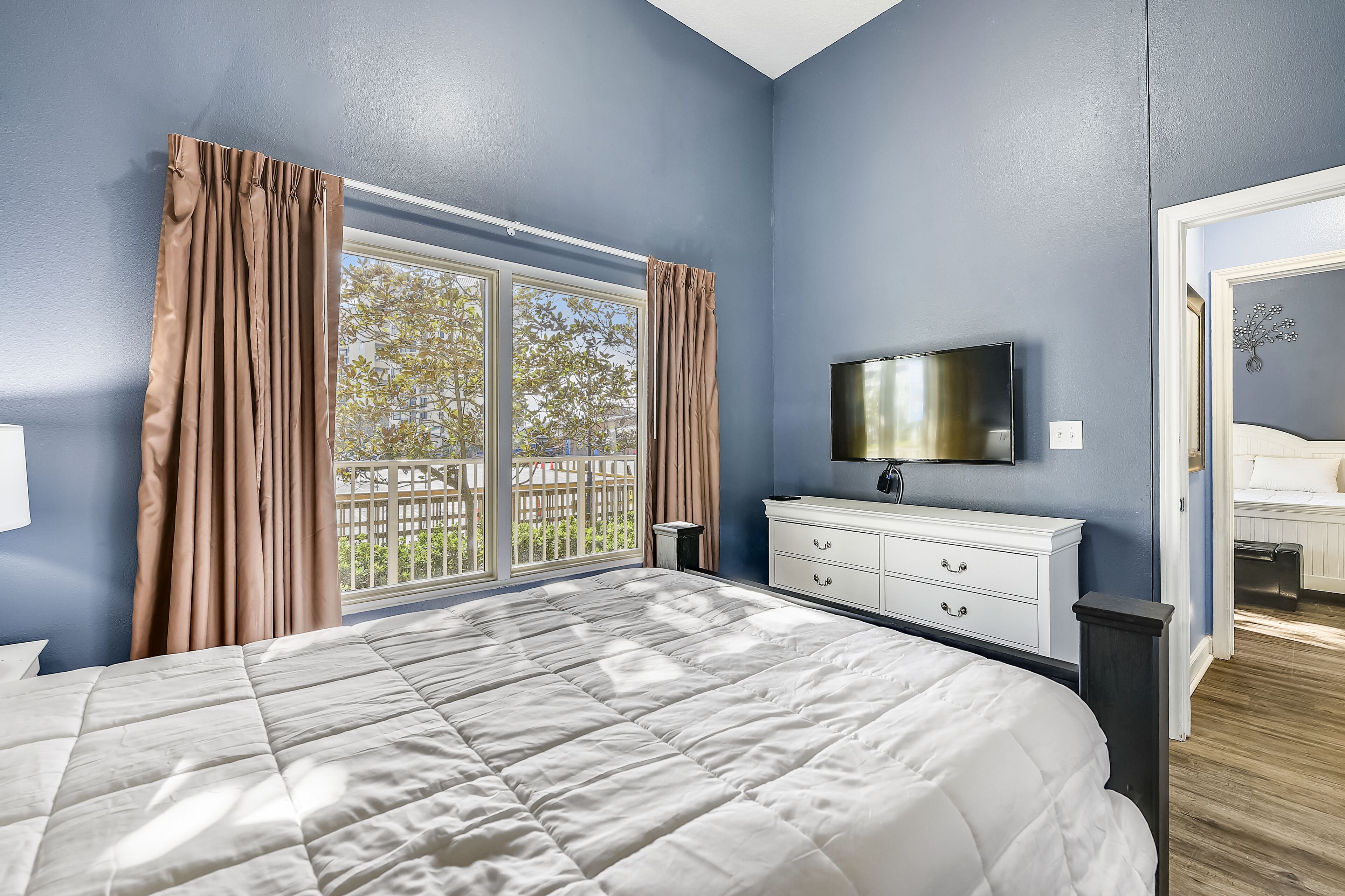 A bedroom with a large bed, a TV mounted on the wall, a dresser, and a window with curtains.