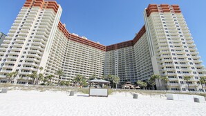 A high-rise hotel with multiple balconies, a beach area with palm trees, and a small pavilion.